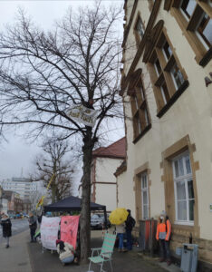 Mahnwache vor dem Gericht in Grevenbroich mit Menschen, Transparenten und Pavillion. Im Baum ist oben eine Person mit Transparent "Stop all fossil fuels"