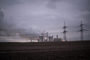 Rauchendes Kohlekraftwerk, davor Strommasten, im Hintergrund wolkenverhangener Himmel