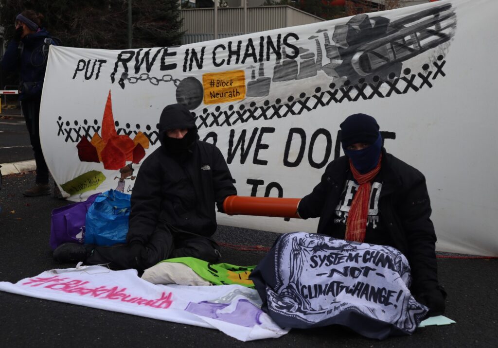 Zwei Menschen sitzen vor Transparent "Put rwe in chains - so we dont hae to" mit Rohr zwischen sich, beide haben ein Arm in einem Rohr