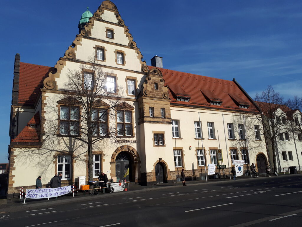 Gerichtsgebäude, gelbes Haus mit Aufschrift "Amtsgericht", davor Mahnwache mit Transparenten, blauer Himmel oben drüber