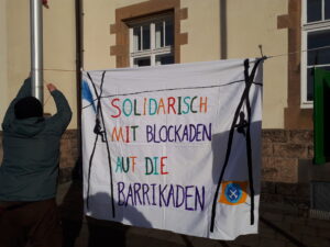 Mensch hängt Transparent zwischen Laternenmasten auf, darauf steht "Solidarisch mit Blockaden auf die Barrikaden"