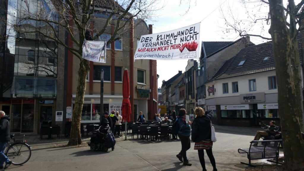 Banner über dem Markplatz "Der Klimawandel verhandelt nicht - wir auch nicht mehr - Ciao Grevenbroich", unten drunter sitzen Menschen im Eiscafé, einige stehen herum.