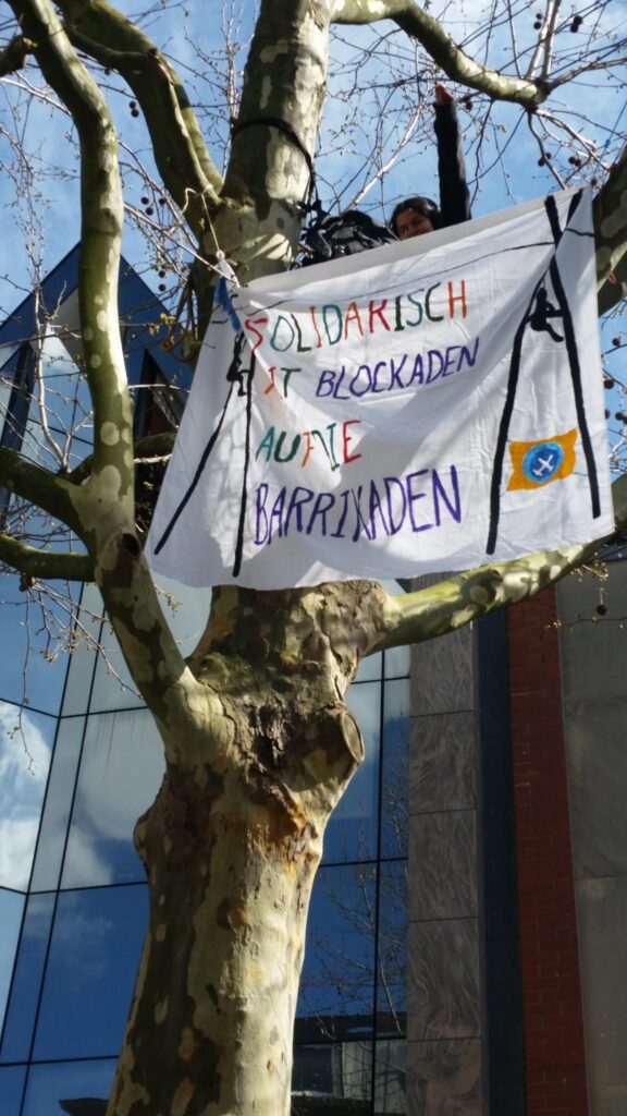 Banner im Baum, Person sitzt darüber, auf dem Banner steht "Solidarisch mit Blockaden auf die Barrikaden"
