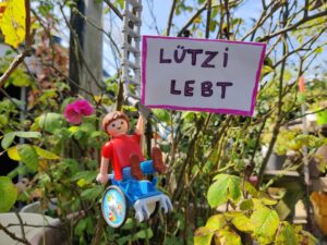 Im Gebüsch hängt eine Playmobilfigur im Rollstuhl und hält ein Schild "Lützi lebt" hoch.