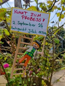 An einer Leiter im Gebüsch hängt eine Playmobilfigur mit einem Schild "Kommt zum Prozess - 2. September 24 Amtsgericht Jülich 11.30 Uhr"