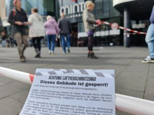 Im Hintergrund Einkaufszentrum "minto", davor verschwommen Menschen, eine Person hat Absperrband in der Hand. Im Vordergrund Absperrband, davor ein Flugblatt mit der Aufschrift "Achtung Luftverschmutzung" Dieses Gebäude ist gesperrt.