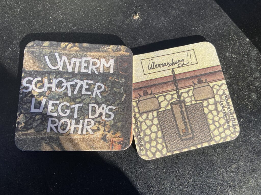 Bierdeckel. Vorderseite zu sehen: "Unterm Schotter liegt das Rohr"-Schriftzug auf Gleisbett. Auf der Rückseite steht "Überraschung" und es ist die Skizee einer Ankettvorrichtung unter einem Gleis zu sehen, daneben steht #blockNeurath und #unfreiwilligeFeuerwehr