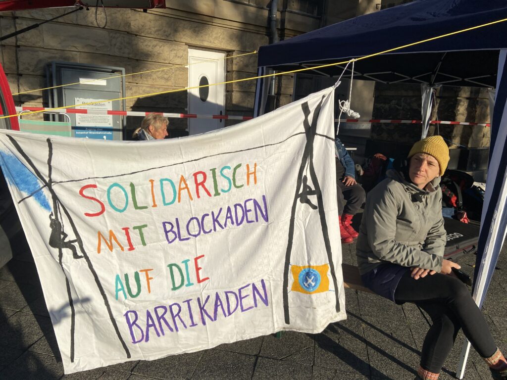 Links im Bild ein Transparent mit aufgemalten Tripods und Aufschrift "Solidarisch mit Blockaden auf die Barrikaden", rechts im Bild sitzt eine Person in der Sonne unterm Pavillion, im Hintergrund ein Gebäude