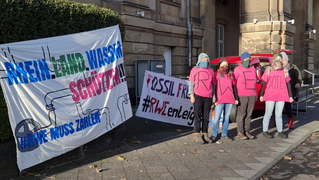 Es ist im Hintergrund ein großes Gebäude zu sehen, davor zwei Transparente mit der Aufschrift "Rhein.Land.Wasser schützen - RWE muss zahlen" und "Fossil free future #RWE enteignen". Davor stehen vier bunt vermummte Personen in pinken T-Shirts, auf denen zusammen gelesen werden kann "RWE tötet".