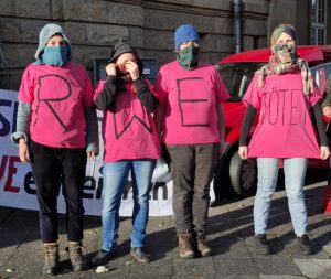 Vier Menschen stehen nebeneinander, vermummt und mit pinken T-Shirts an, auf den T-Schirts der ersten drei Personen stehen die Buchstaben R, W und E, auf dem T-Shirt der letzten Person "TÖTET!".