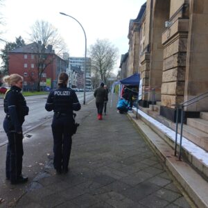 Zwei Polizistinnen stehen im Vordergrund und beobachten Menschen, die damit beschäftigt sind auf die Stufen eines großen Gebäudes weiße Plakate zu legen.