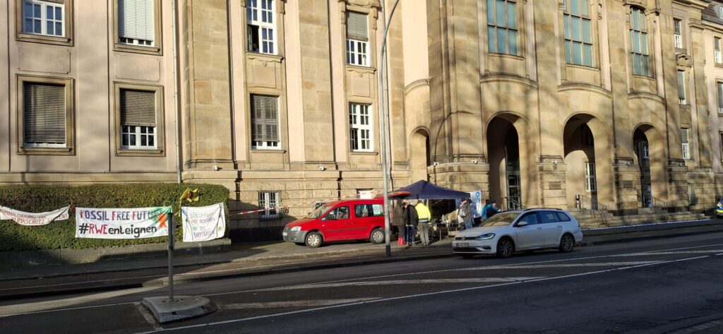 Im Hintergrund ein großes Gebäude, vorne eine Straße. Vor dem Gebäude auf dem Bürgerstieg sind Menschen, ein Auto, ein Pavillion und an einer Hecke hängen Banner mit Aufschriften wie "Fossil free future #RWE enteignen"