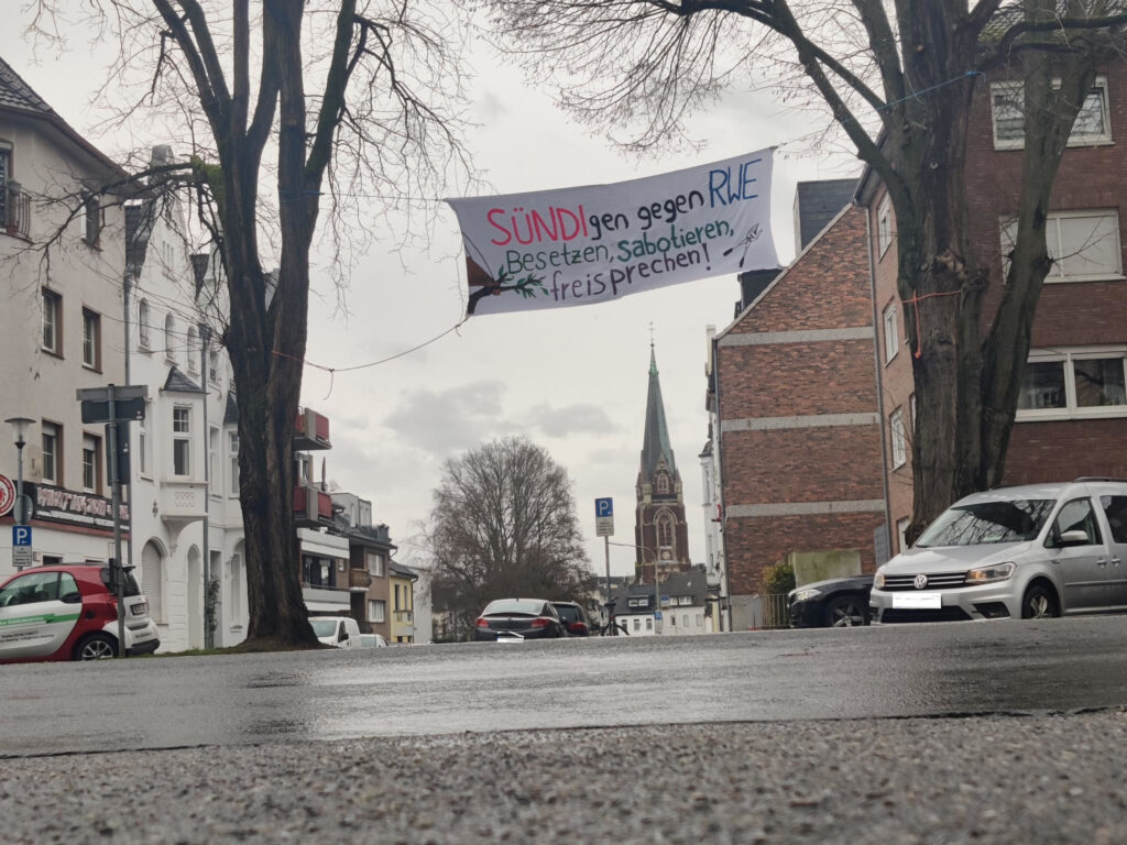 Banner "SÜNDIgen gegen RWE. Besetzen, sabotieren, freisprechen" zwischen zwei Bäumen aufgehangen. Im HIntergrund ein Kirchturm, im Vordergrund die Straße.