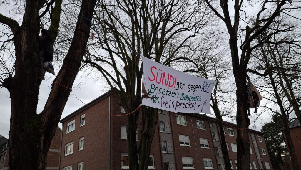 Banner "SÜNDIgen gegen RWE. Besetzen, sabotieren, freisprechen" zwischen mehreren Bäumen aufgehangen. In den Bäumen hängen zwei Menschen, im Hintergrund ist ein Hochhaus zu sehen.