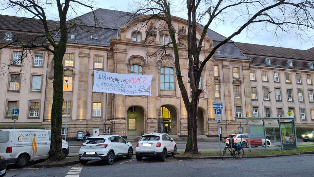 Das große Sandsteingebäude des Landgerichts Mönchengladbach ist im Hintergrund zu sehen, davor ein Transparent zwischen zwei Bäumen, welches allerdings nur von hinten zu sehen ist. Ein paar Autos sind geparkt.