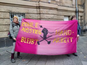 Zwei Menschen halten ein pinkes Transparent, in der Mitte ist eine Faust mit gezogenem Stecker, darunter gekreuzte Hämmer, in gelber Schrift ist zu lesen: "Kohleausstieg bleibt Handarbeit" - im HIntergrund ist ein Gebäude zu sehen.
