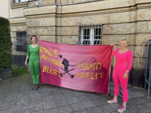 Zwei Menschen in einem grünen bzw. pinken Ganzkörperanzug halten ein pinkes Transparent, in der Mitte ist eine Faust mit gezogenem Stecker, darunter gekreuzte Hämmer, in gelber Schrift ist zu lesen: "Kohleausstieg bleibt Handarbeit" - im HIntergrund ist ein Gebäude zu sehen.