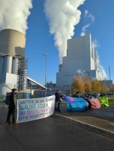 Mehrere Menschen stehen mit Transparent vor einem rauchenden Kohlekraftwerk auf der Einfahrt.