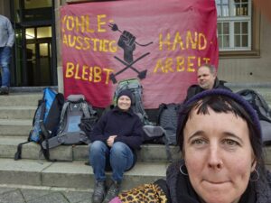 Drei Menschen sitzen mit Rucksäcken auf den Stufen des Gerichts vor einem Transparent.