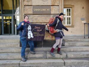 Zwei Menschen bewegen sich auf Treppenstufen vor einer Säule mit einem Messingschild "Landgericht" mit einem Transparent mit aufgemaltem Schneemann und der Aufschrift "Save Olaf - smash capitalism"