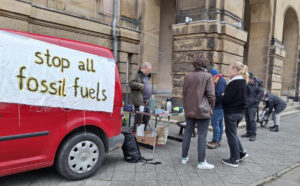 An einem roten Auto links im Bild hängt ein Banner mit der Aufschrift "stop all fossil fuels", rechts im Bild stehen mehrere Menschen um einen Klapptisch mit Getränken herum, im Hintergrund ist ein Gebäude zu sehen.