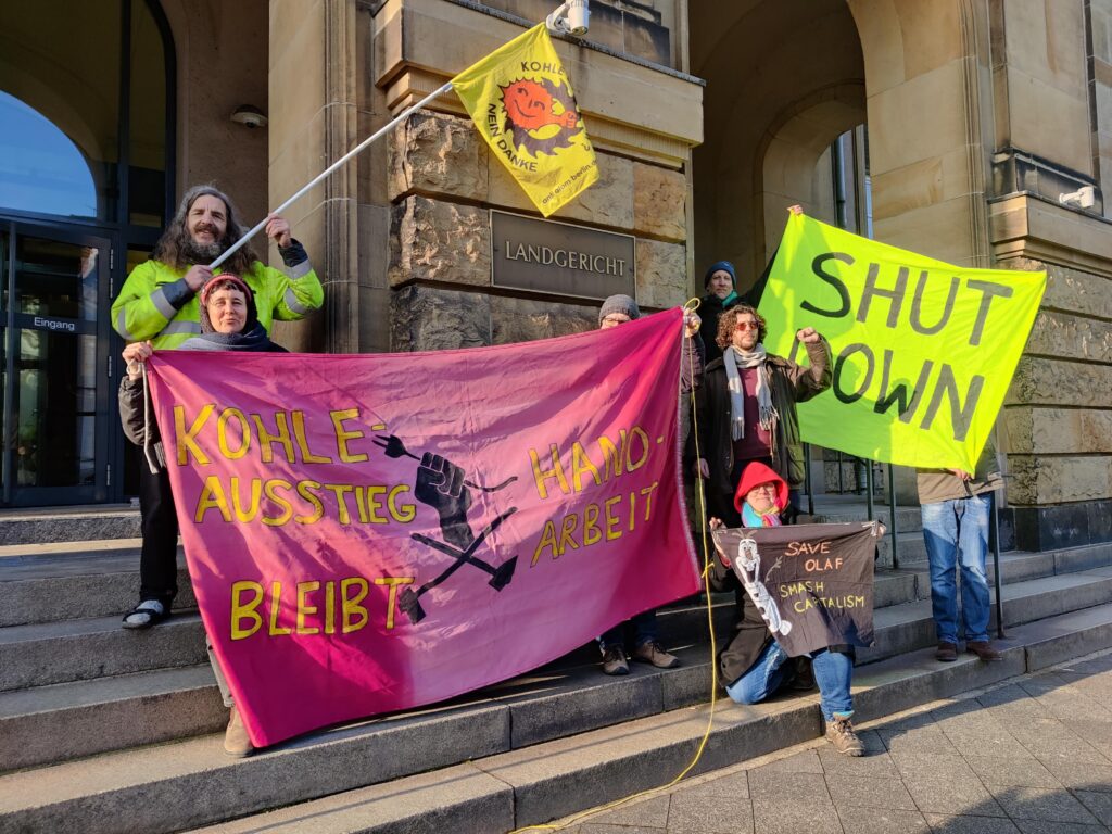 Mehrere Menschen mit Transparenten u.a. "Kohleausstieg bleibt Handarbeit" vor dem Schild "Landgericht" auf Treppenstufen