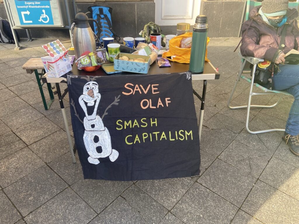 Kleines schwarzes Transparent "Save Olaf - smash Capitalism" mit Schneemensch drauf an einem Tisch mit Teekannen und Essen drauf